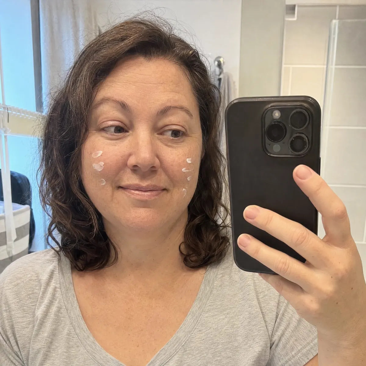 A woman with dabs of white cream on her cheeks takes a selfie in a mirror.