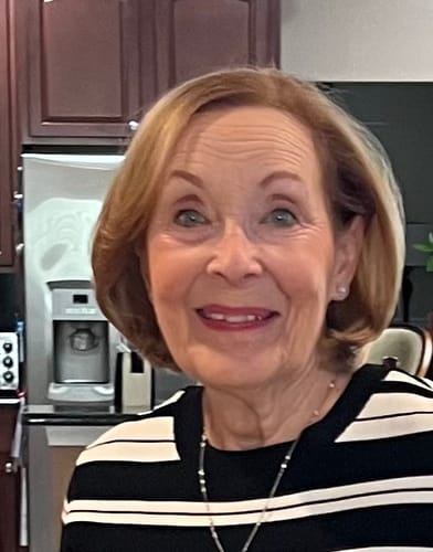 A smiling older woman with short, reddish-blonde hair wearing a black and white striped top.